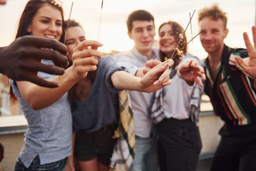 With sparklers in hands. Group of young people in casual clothes have a party at rooftop together at daytime.