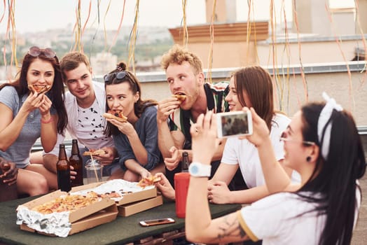 Making photo. Delicious pizza. Group of young people in casual clothes have a party at rooftop together at daytime.