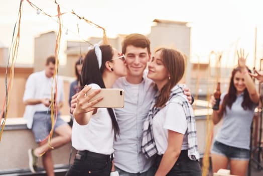 Two girls kissing one man. Taking selfie. Group of young people in casual clothes have a party at rooftop together at daytime.