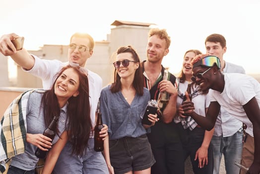 Making selfie by phone. Group of young people in casual clothes have a party at rooftop together at daytime.