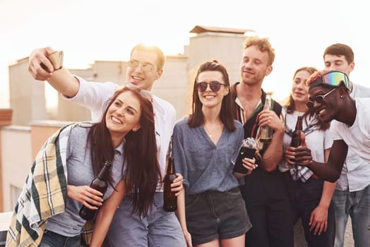 Making selfie by phone. Group of young people in casual clothes have a party at rooftop together at daytime.