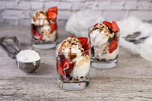 Summer cold dessert - Plombir ice cream scoop with chocolate sauce, cookie crumbs, ripe strawberries, crushed peanut nuts in a glass on a wooden table