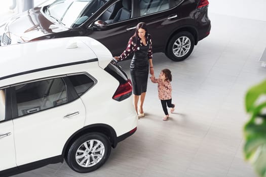 Mother with her daughter walking together near modern automobile indoors.