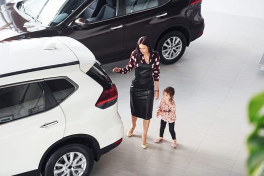 Mother with her daughter walking together near modern automobile indoors.