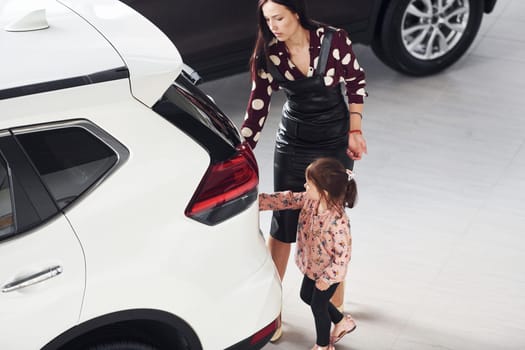 Mother with her daughter walking together near modern automobile indoors.