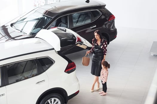 Mother with her daughter walking together near modern automobile indoors.