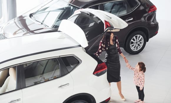 Mother with her daughter walking together near modern automobile indoors.
