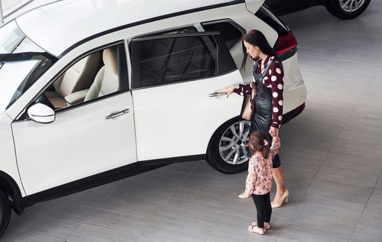 Mother with her daughter walking together near modern automobile indoors.