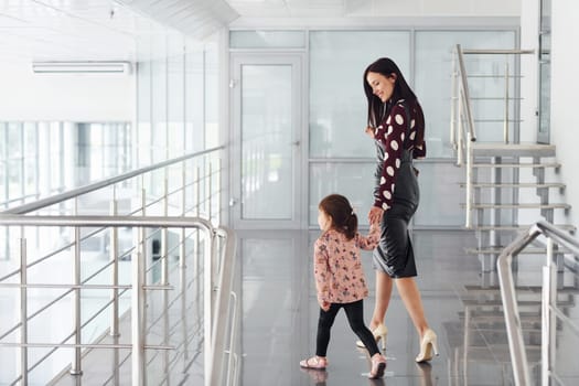 Young mother with her daughter walking together indoors in the office or airport. Having vacation.