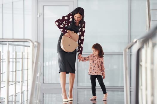 Young mother with her daughter walking together indoors in the office or airport. Having vacation.