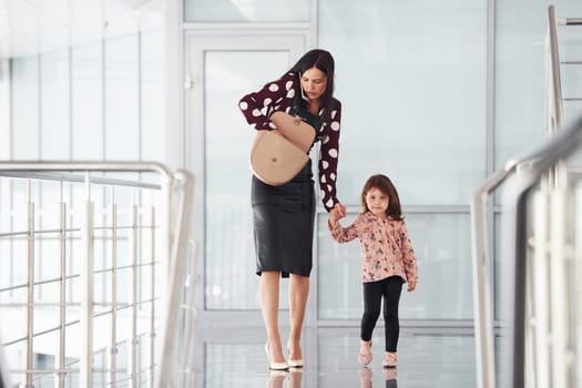 Young mother with her daughter walking together indoors in the office or airport. Having vacation.