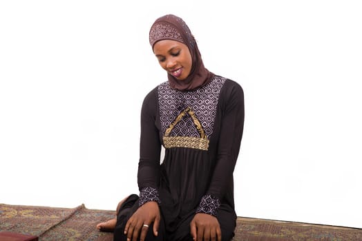 young muslim woman sitting in long dress on mat praying while smiling.