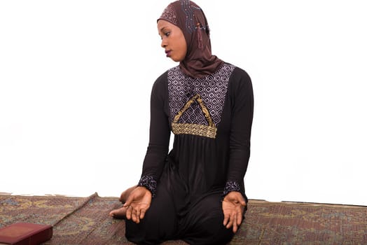 young muslim woman sitting in long dress on mat praying while looking in profile.