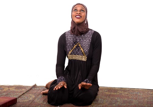 young muslim woman sitting in long dress on mat praying while looking up smiling.