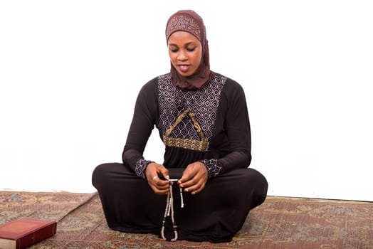 young muslim woman sitting in long dress on mat praying while smiling.