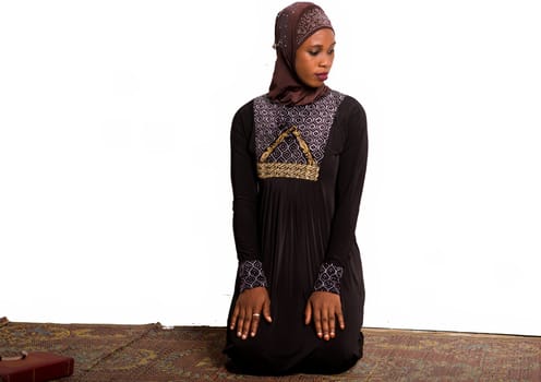 young muslim woman kneeling on a mat praying while looking in profile.