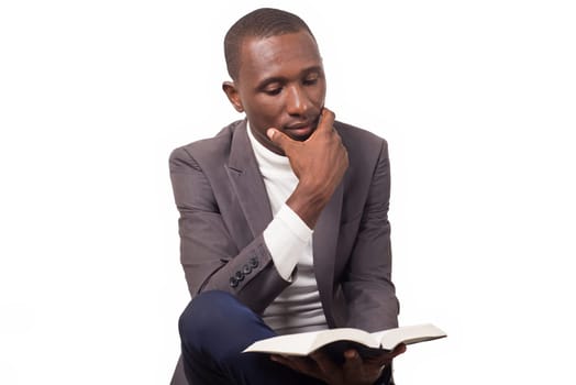 portrait of young pastor sitting cross-legged, hand under chin and reading a Bible he holds in the other hand.