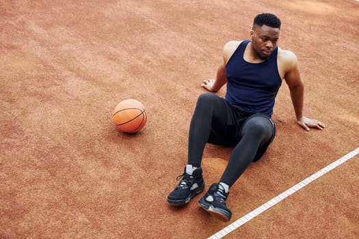 Tired african american man sits on the ground with ball on the court outdoors. Takes a break.