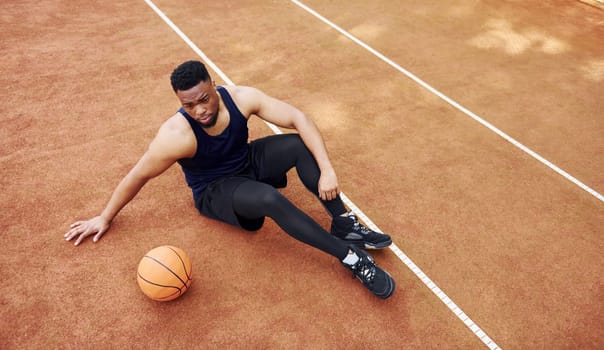 Tired african american man sits on the ground with ball on the court outdoors. Takes a break.