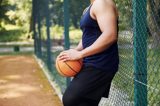 African american man takes a break and leaning on the metal mesh with ball on the court outdoors.