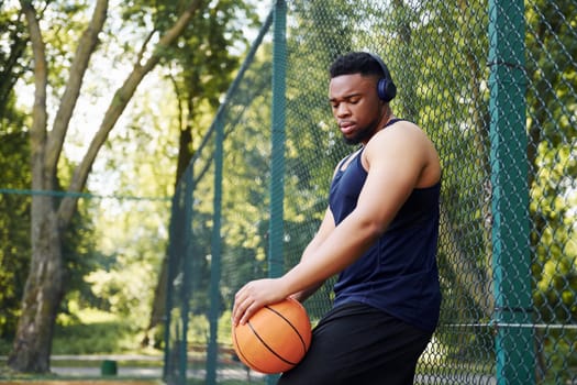 African american man with wireless headphones takes a break and leaning on the metal mesh with ball on the court outdoors.