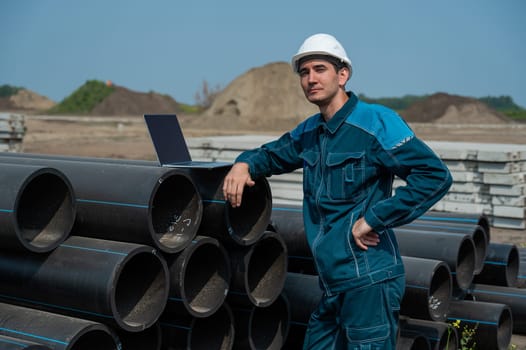 Caucasian male builder in a hard hat stands near the pipes and uses a laptop at a construction site