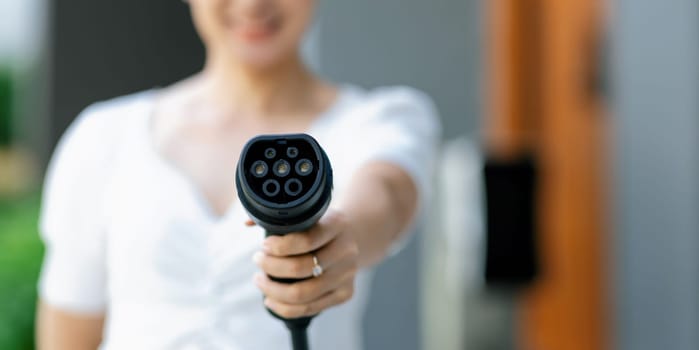 Focus hand holding and pointing EV charger plug for electric vehicle at camera with blur woman in background from home charging station as progressive idea of alternative energy for ecology concern.