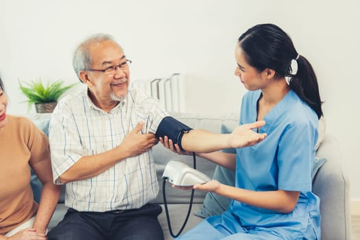 An elderly man having a blood pressure check by his personal caregiver with his wife sitting next to him in their home.