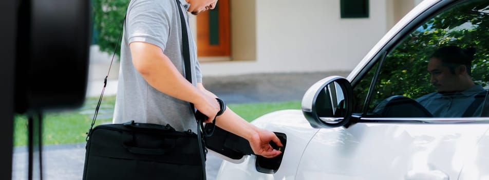 Progressive asian man install cable plug to his electric car with home charging station in the backyard. Concept use of electric vehicles in a progressive lifestyle contributes to clean environment.