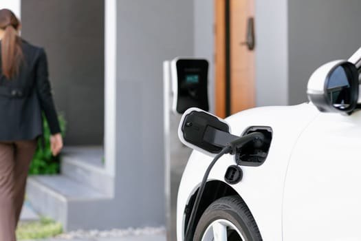 Focus image of electric vehicle recharging battery at home charging station with blurred woman walking in the background. Progressive concept of green energy technology applied in daily lifestyle.