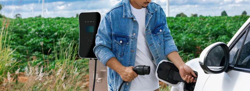 Progressive man with his electric car, EV car recharging energy from charging station on green field with wind turbine as concept of future sustainable energy. Electric vehicle with energy generator.