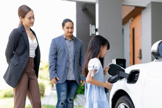 Progressive young parents and daughter with electric vehicle and home charging station. Green and clean energy from electric vehicles for healthy environment. Eco power from renewable source at home.