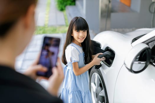 Progressive mother using smartphone take focus photo of daughter recharge EV car. Image of asian girl insert electric charger from home charging station to EV car on phone screen with blur background.