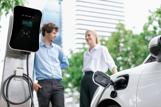 Focus parking-electric car connected to public charging station with blur progressive businesspeople holding coffee, residential building apartment and condo background for eco-friendly concept.