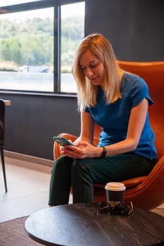 positive blonde businesswoman holding a mobile phone, happy to receive a message from her best friend or colleague, sitting in a cozy cafeteria at the table, High quality photo