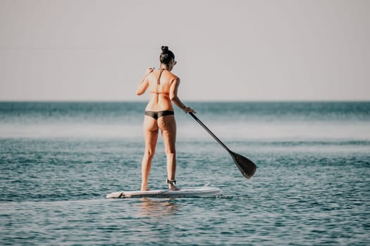 Sea woman sup. Silhouette of happy middle aged woman in rainbow bikini, surfing on SUP board, confident paddling through water surface. Idyllic sunset. Active lifestyle at sea or river