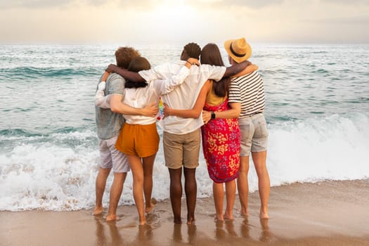 Rear view of multiracial friends embracing together looking at the ocean relaxing during vacation trip. Friendship concept.