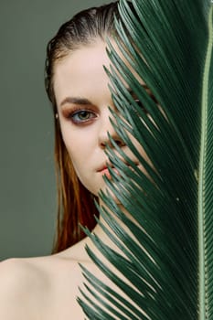 a sophisticated, elegant woman with evening makeup, stands with a green palm leaf, covering part of her face with it. Vertical photo without retouching. High quality photo
