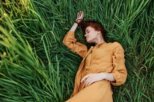 a close horizontal photo of a pleasant woman in a long orange dress resting lying in the tall grass with her eyes closed in sunny weather. High quality photo