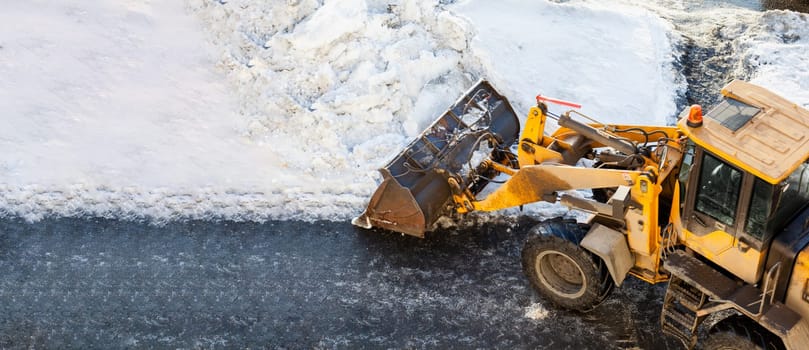 Orange tractor cleans up snow from the road and loads it into the truck. Cleaning of roads in the city from snow in winter. Snow removal after snowfall and blizzards.