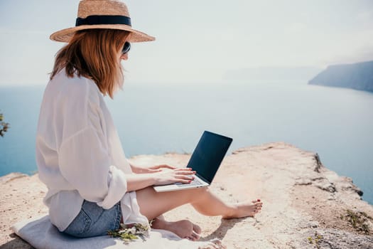 Successful business woman in yellow hat working on laptop by the sea. Pretty lady typing on computer at summer day outdoors. Freelance, travel and holidays concept.