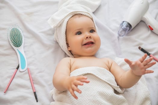 beautiful baby with a white towel on her head lies near the hairdryer, comb and cosmetics