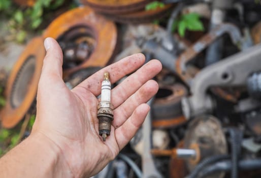 old spark plug in hand near a pile of rusty spare parts in a landfill.