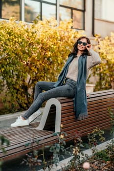 Woman wearing glasses, beige sweater, jeans, white sneakers. Happy lifestyle in the city with autumn trees in view.