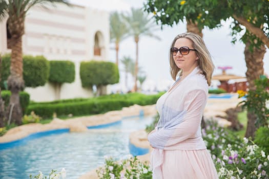 blonde in sunglasses and shawls standing in the garden with pool.