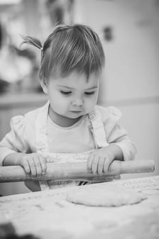 Adorable baby in an apron is rolling out the dough in the kitchen.