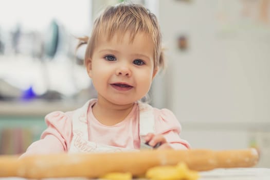 Adorable baby in an apron is rolling out the dough in the kitchen.