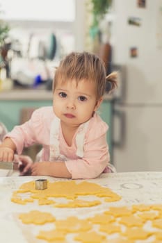 Adorable baby in an apron makes cookies from dough in the kitchen.