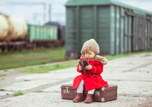 charming baby in a red coat with a suitcase is waiting for a train on the platform