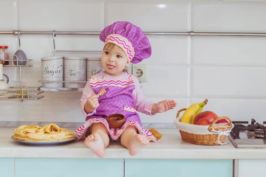 Adorable baby in an apron and chef's cap eating jam in the kitchen.